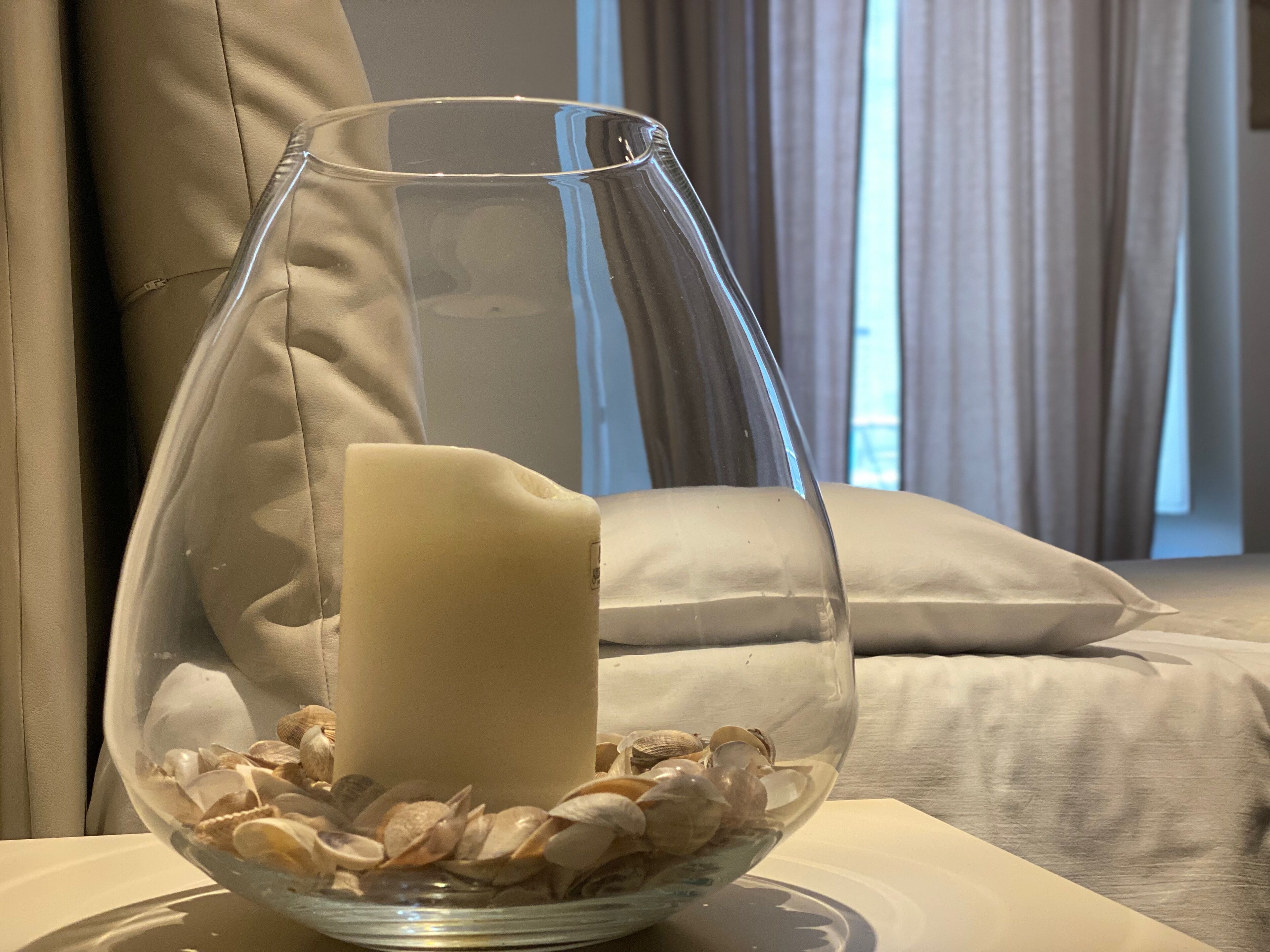 Un tazón de vidrio transparente con una vela blanca y conchas marinas, sobre una mesa con una cama y cortinas al fondo.