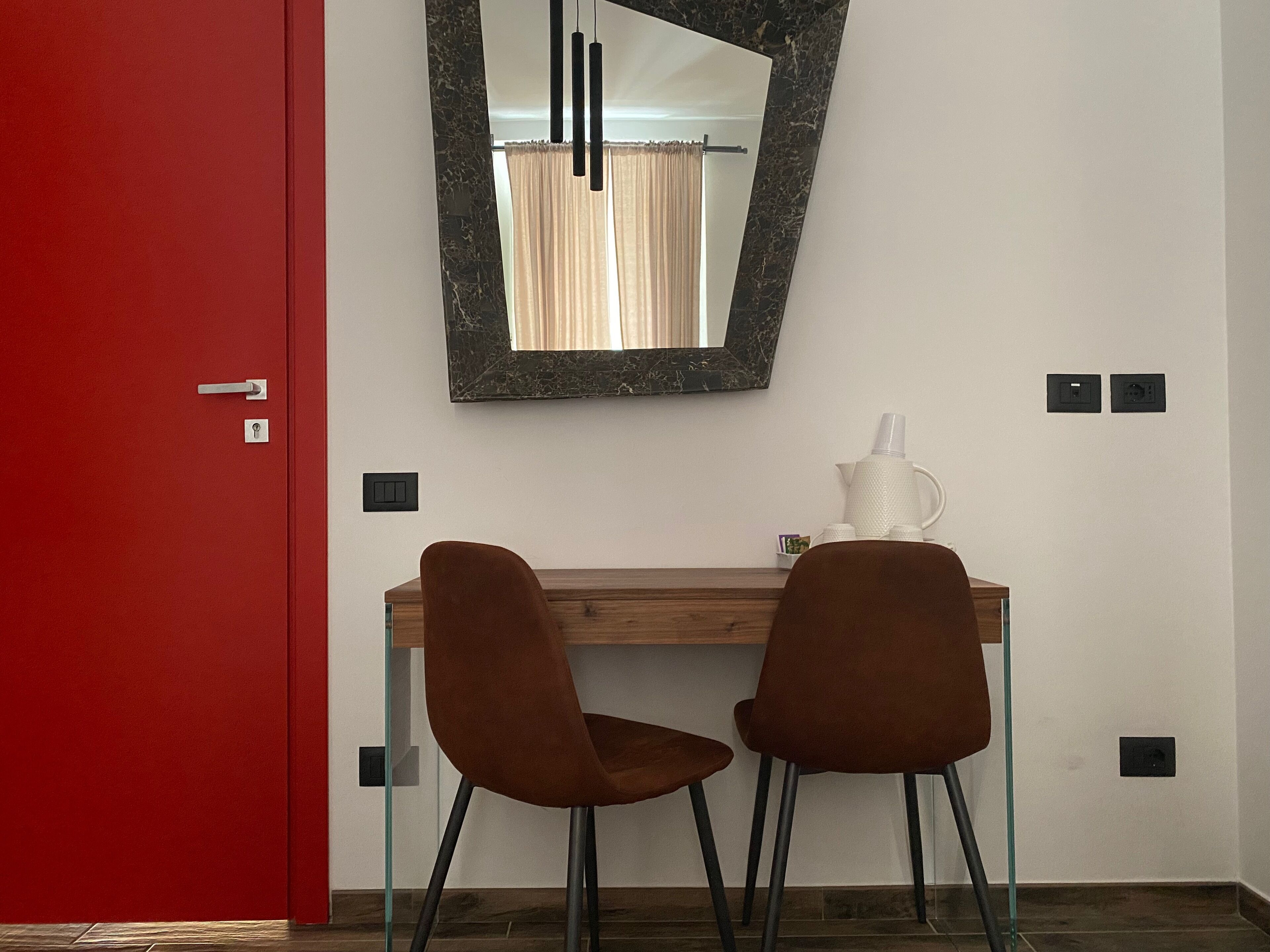 Una habitación con una puerta roja, un escritorio de madera, dos sillas marrones, una jarra blanca y un espejo con marco de mármol.
