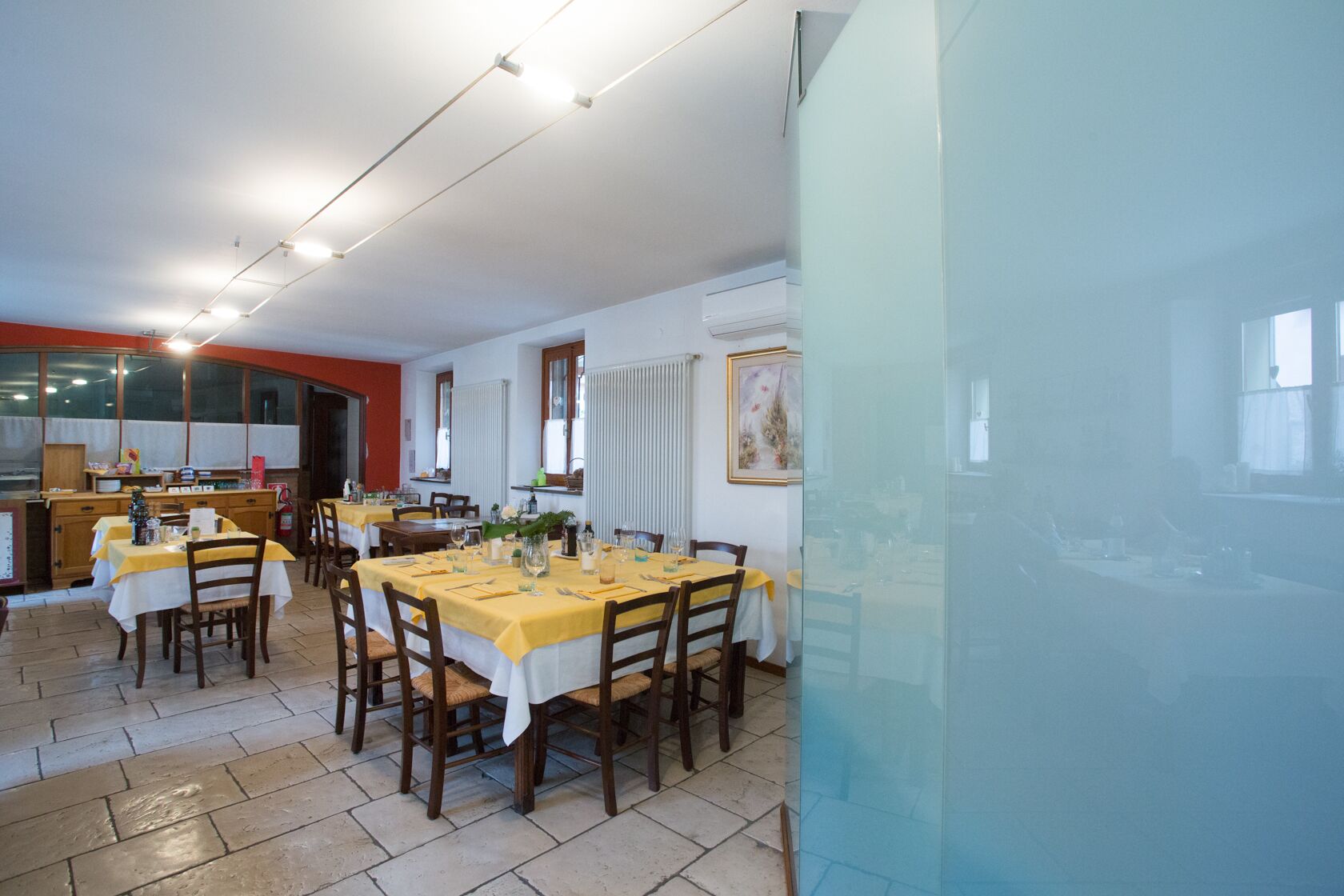 Un espace repas avec plusieurs tables dressées pour les repas, un coin cuisine et une grande fenêtre qui laisse entrer la lumière naturelle.