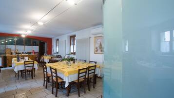 Un espace repas avec plusieurs tables dressées pour les repas, un coin cuisine et une grande fenêtre qui laisse entrer la lumière naturelle.