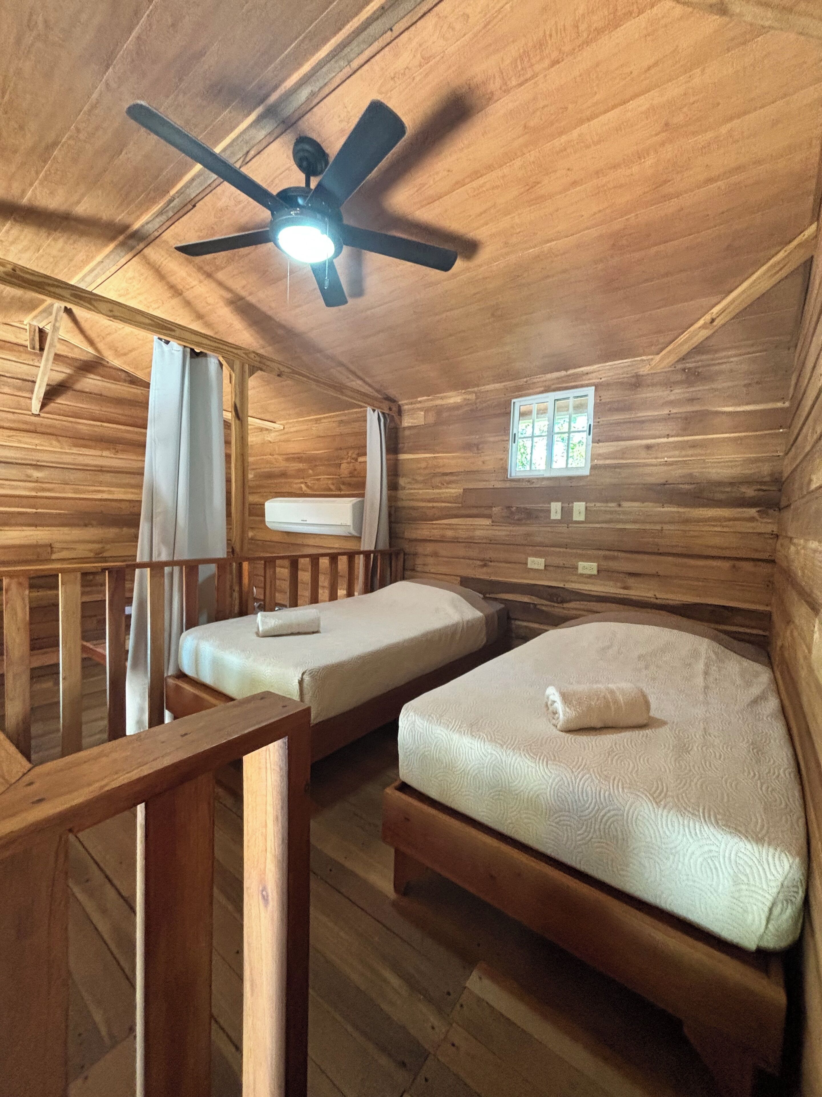 Una cabaña de madera con dos camas, un ventilador de techo, una ventana y un equipo de aire acondicionado.