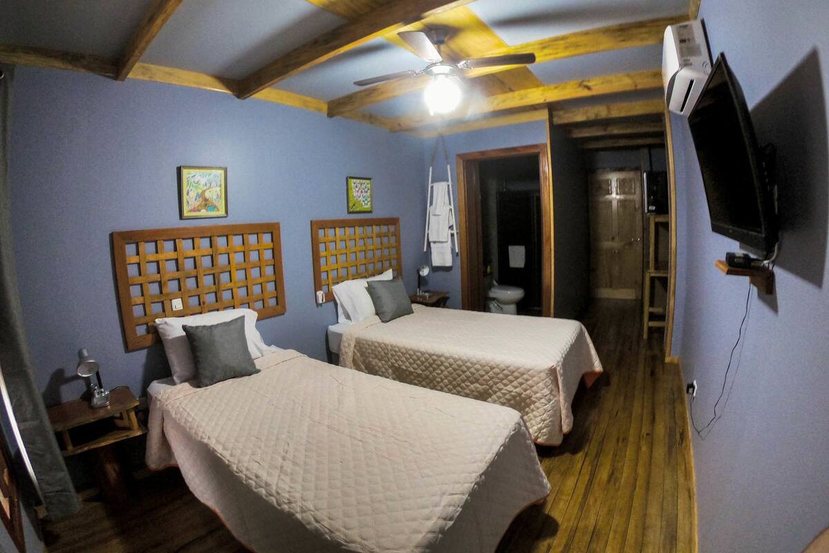 Une chambre avec deux lits, des têtes de lit en bois, un ventilateur de plafond, un téléviseur fixé au mur et une salle de bain visible à travers une porte ouverte.