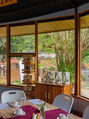 Un coin repas avec une table dressée pour quatre personnes, un espace dédié au café et une vue sur un jardin agrémenté d’arbres et d’une clôture.