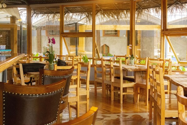 Un restaurante con mesas y sillas de madera, ventanas amplias y un techo de paja.