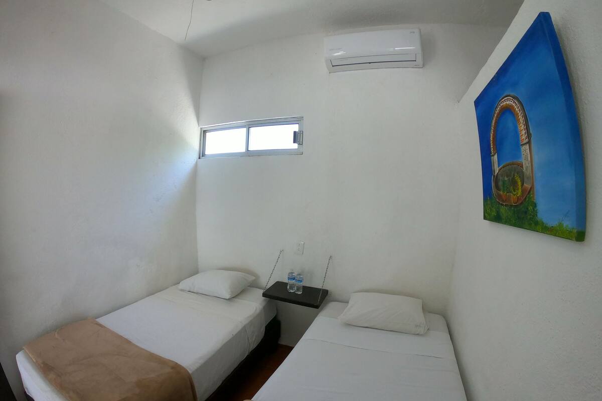 Una habitación pequeña y limpia con dos camas individuales, una ventana, un equipo de aire acondicionado y un cuadro en la pared.