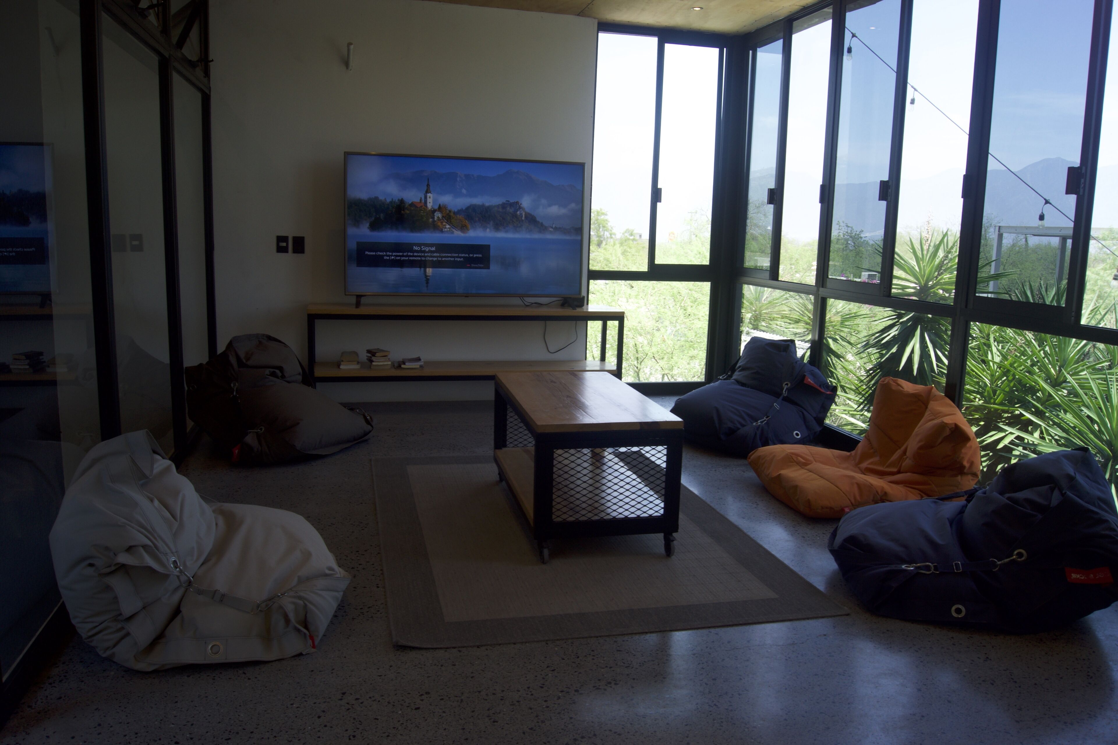 A modern living room with a large TV, comfortable seating, and a view of greenery through large windows.