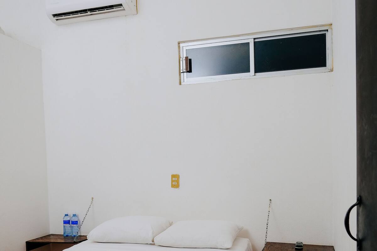 Una habitación pequeña y limpia con cama, aire acondicionado, ventana y mesita de noche con una botella de agua.