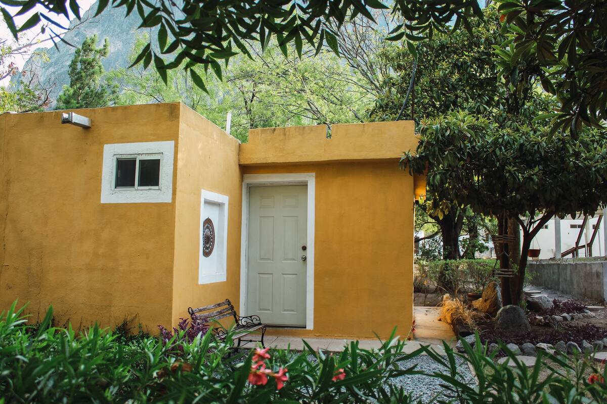 Pequeño edificio amarillo con puerta y ventana blancas, rodeado de vegetación y un banco.