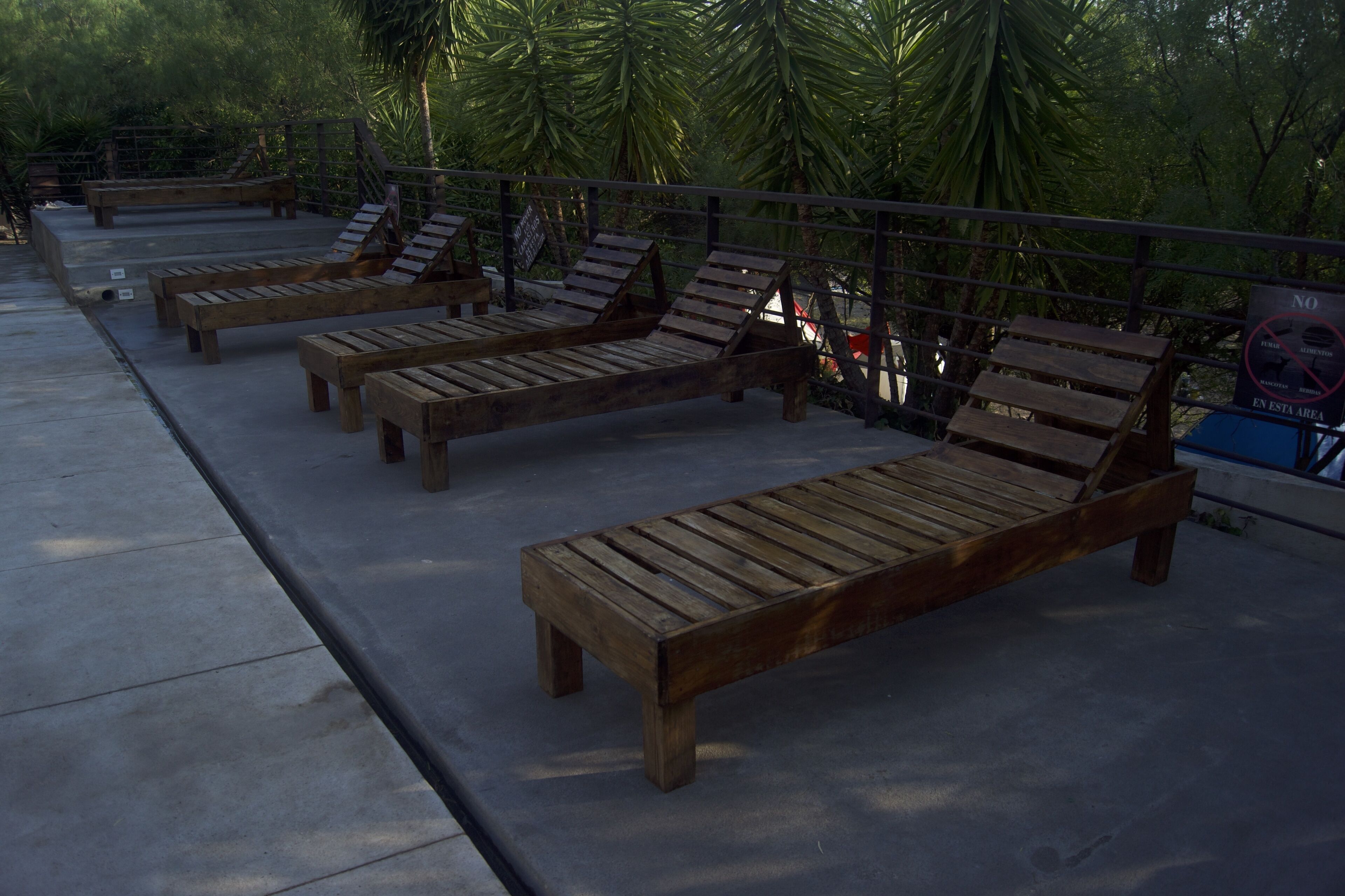 Wooden lounge chairs arranged on a concrete deck with a metal railing, surrounded by greenery.