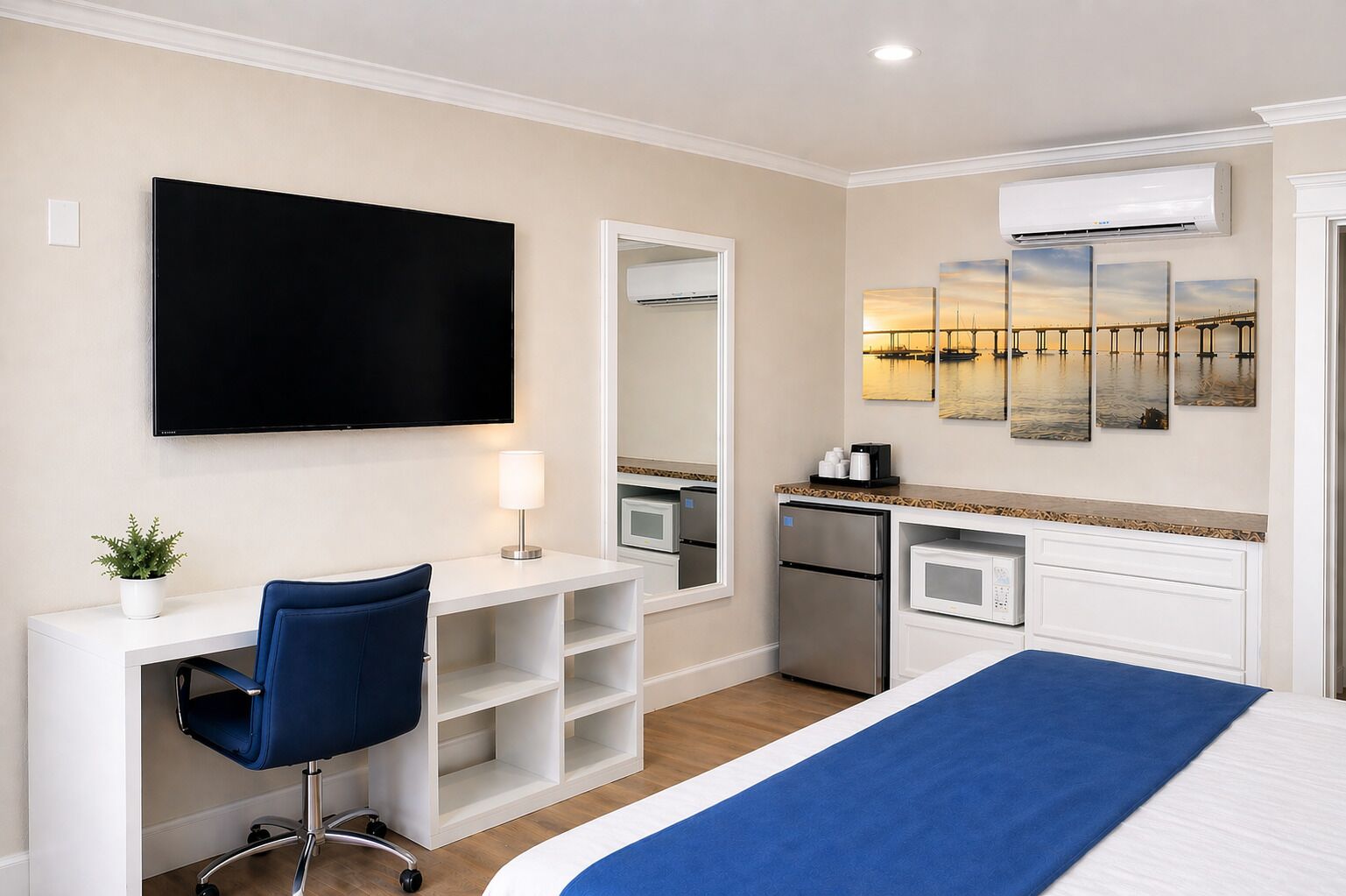 A hotel room with a bed, a desk with a chair, a flat-screen TV, a microwave, a refrigerator, and a large mirror.