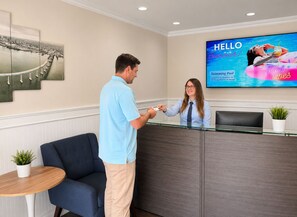A reception area with a receptionist, a man handing over a card, a large TV screen displaying a pool advertisement, and a framed cityscape artwork. - Coronado Inn (Coronado)
