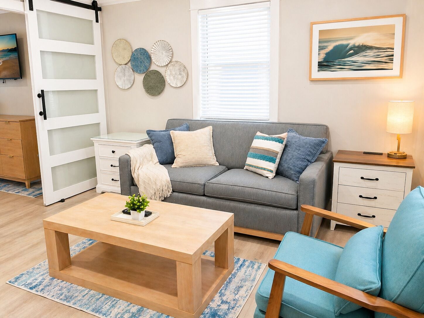 A modern living room with a grey sofa, blue and white cushions, a wooden coffee table, a white side table, and a wall-mounted picture of a wave.