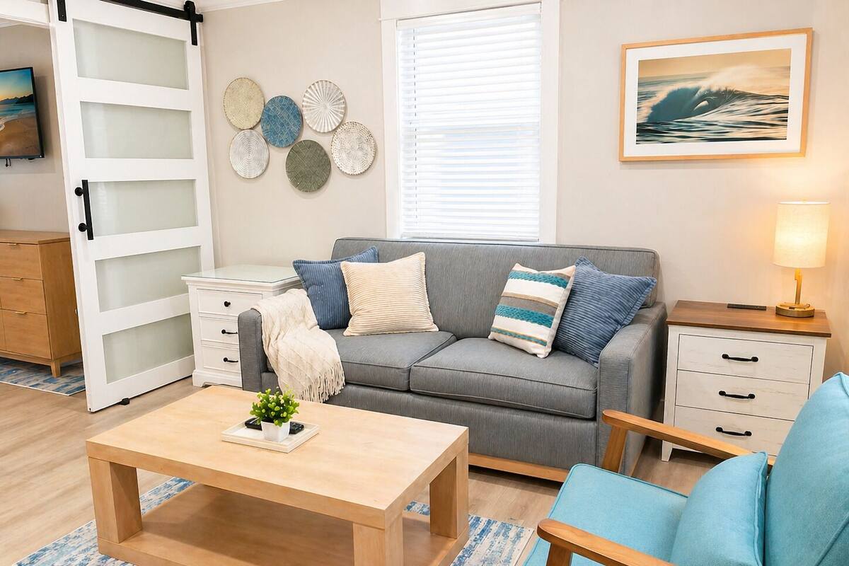 Un salon moderne avec un canapé gris, des coussins bleus et blancs, une table basse en bois, une petite table d’appoint blanche et un tableau représentant une vague, accroché au mur.