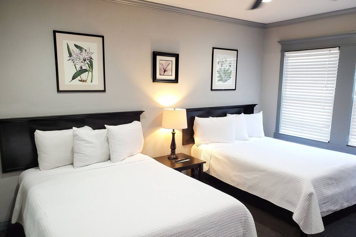 Une chambre d’hôtel avec deux lits, une table de chevet avec une lampe et des reproductions encadrées de motifs botaniques accrochées au mur.