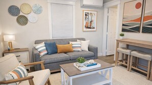 A living room with a grey sofa, a white coffee table, a wooden side table, and a chair. There is a painting on the wall and a window with blinds. - Coronado Inn (Coronado)