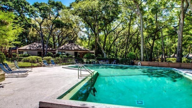 Una piscina con sillones de descanso, rodeada de árboles y un edificio con techo de paja.