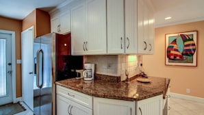 Una cocina con gabinetes blancos, encimera de granito, refrigerador y cafetera.