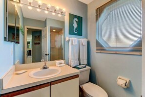 A bathroom with a sink, mirror, and toilet. There is a towel rack with towels, a seahorse painting on the wall, and a window with blinds. - 212-sailmaker (Fernandina Beach)