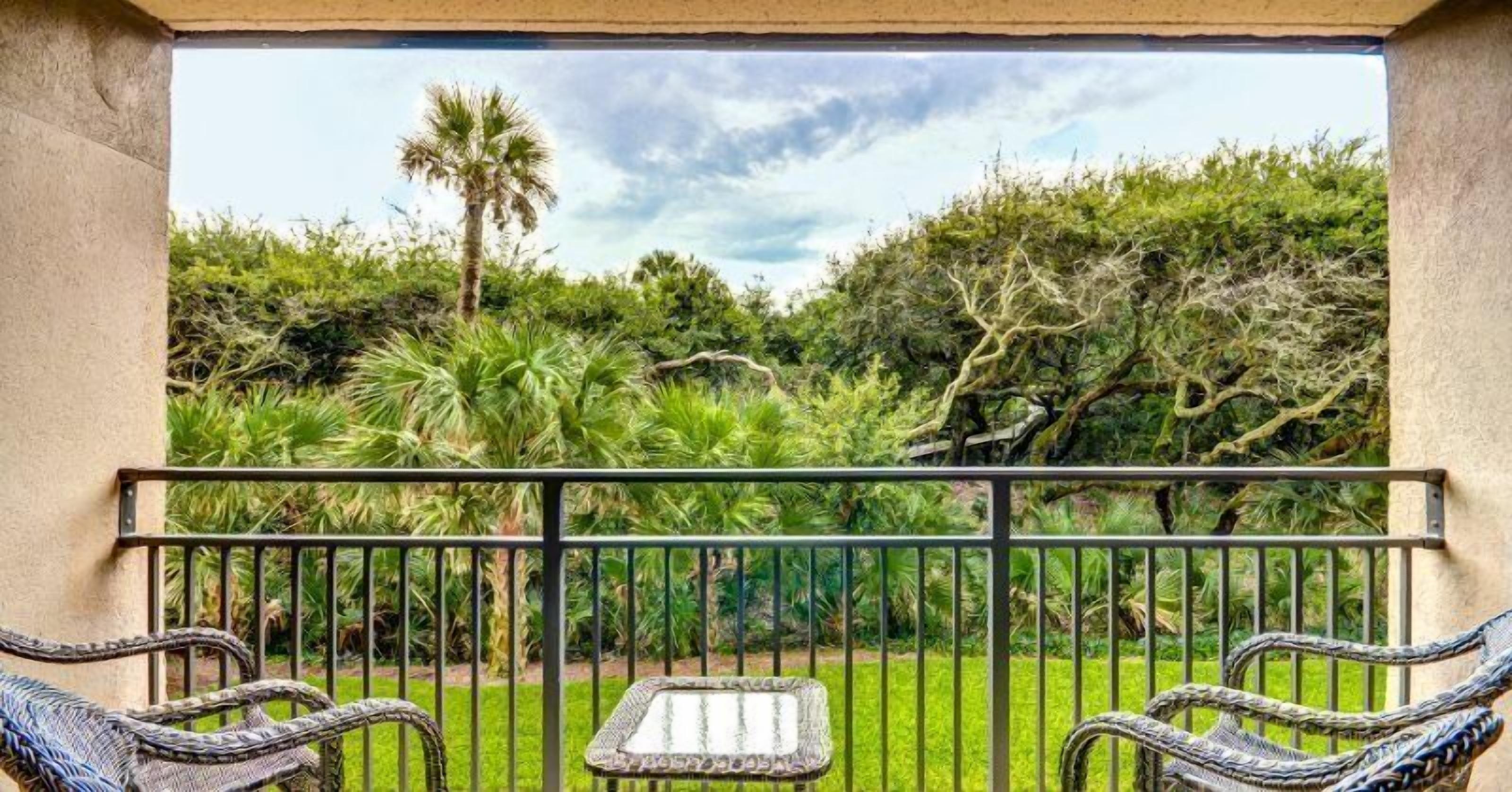 A balcony with wicker chairs and a small table, overlooking a lush garden with various plants and a palm tree.