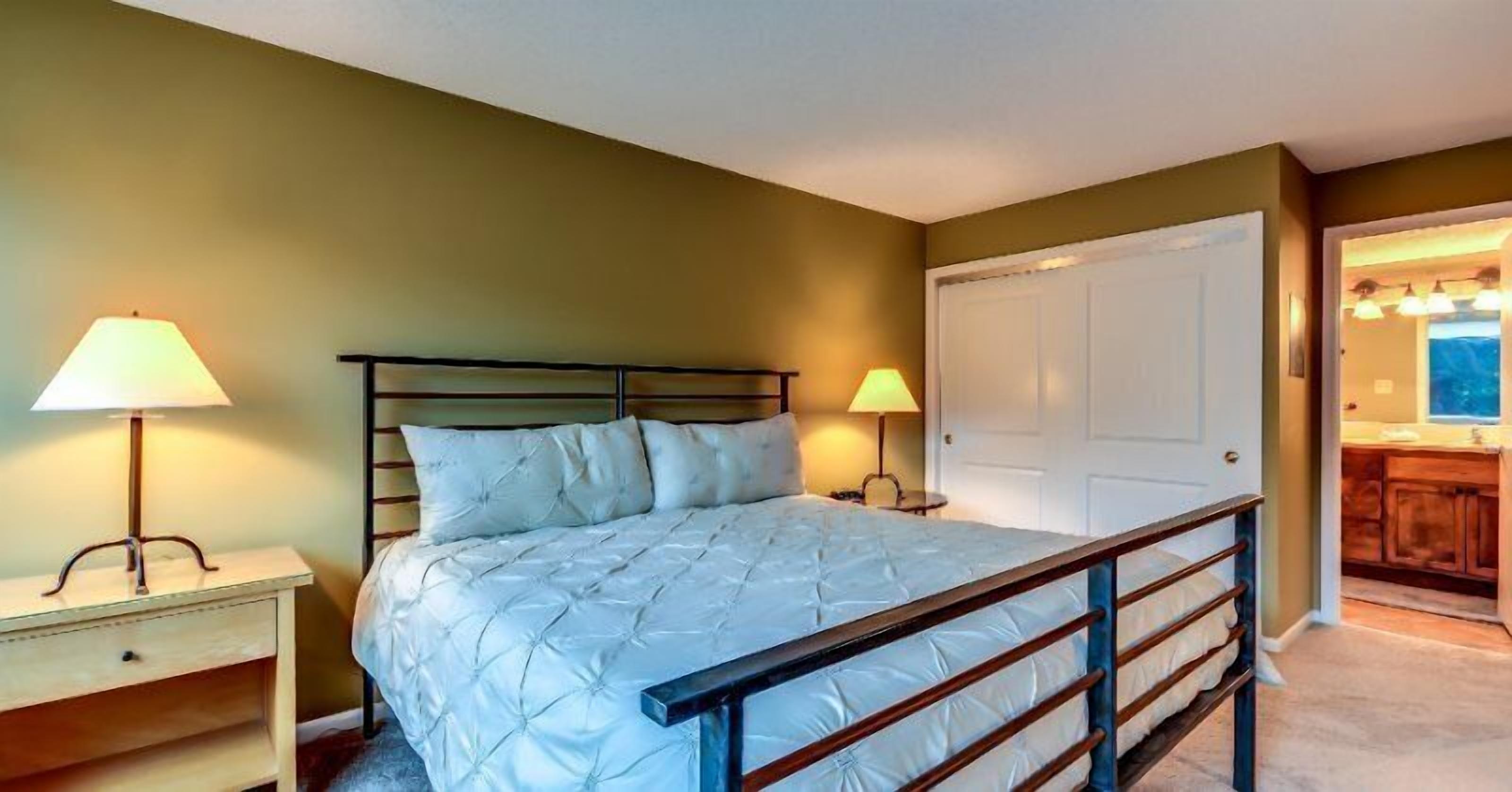 A bedroom with a bed, two bedside lamps, a wooden nightstand, a door, and a view of a bathroom with a sink and mirror.