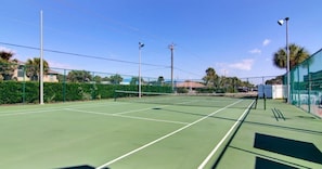 An outdoor tennis court with a net, surrounded by a green fence, palm trees, and a clear sky.