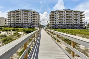 Une promenade en bois mène à un immeuble d’appartements situé en bord de mer, doté de nombreux balcons.