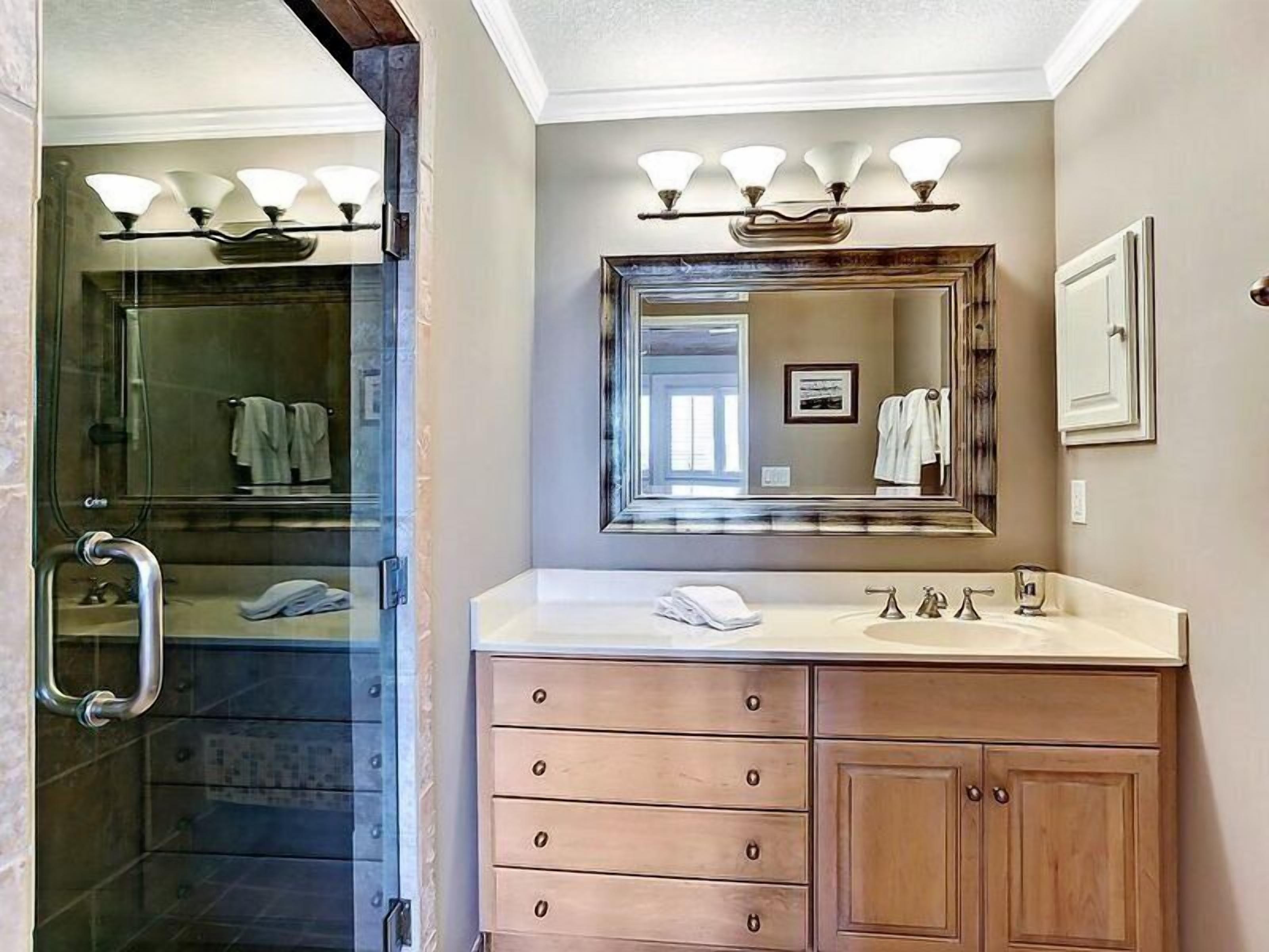A bathroom with a glass shower enclosure, a vanity with a mirror and lights, and wooden cabinets.