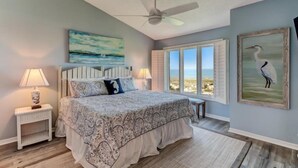 Un dormitorio con cama, ventilador de techo, mesita de noche con lámpara, ventana con vista a la playa y un cuadro de un pájaro en la pared.