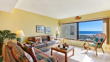 Uma sala de estar com sofá, mesa de centro e uma poltrona. Há uma janela grande com vista para a praia.