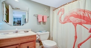 Een badkamer met een houten wastafelmeubel, een wit toilet, een zeester decoratie en een roze flamingo douchegordijn.