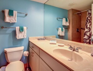A bathroom with a blue wall, white toilet, beige sink, and a shower with a patterned curtain. - 1031-captains-court (Fernandina Beach)