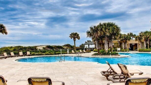 A pool area with lounge chairs, palm trees, and a building in the background. - 1637-38-sea-dunes (Fernandina Beach)