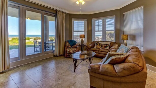 A spacious living room with a large sliding glass door leading to a balcony, a brown leather sofa, a small round coffee table, and a ceiling fan. - 5-ocean-place (Fernandina Beach)