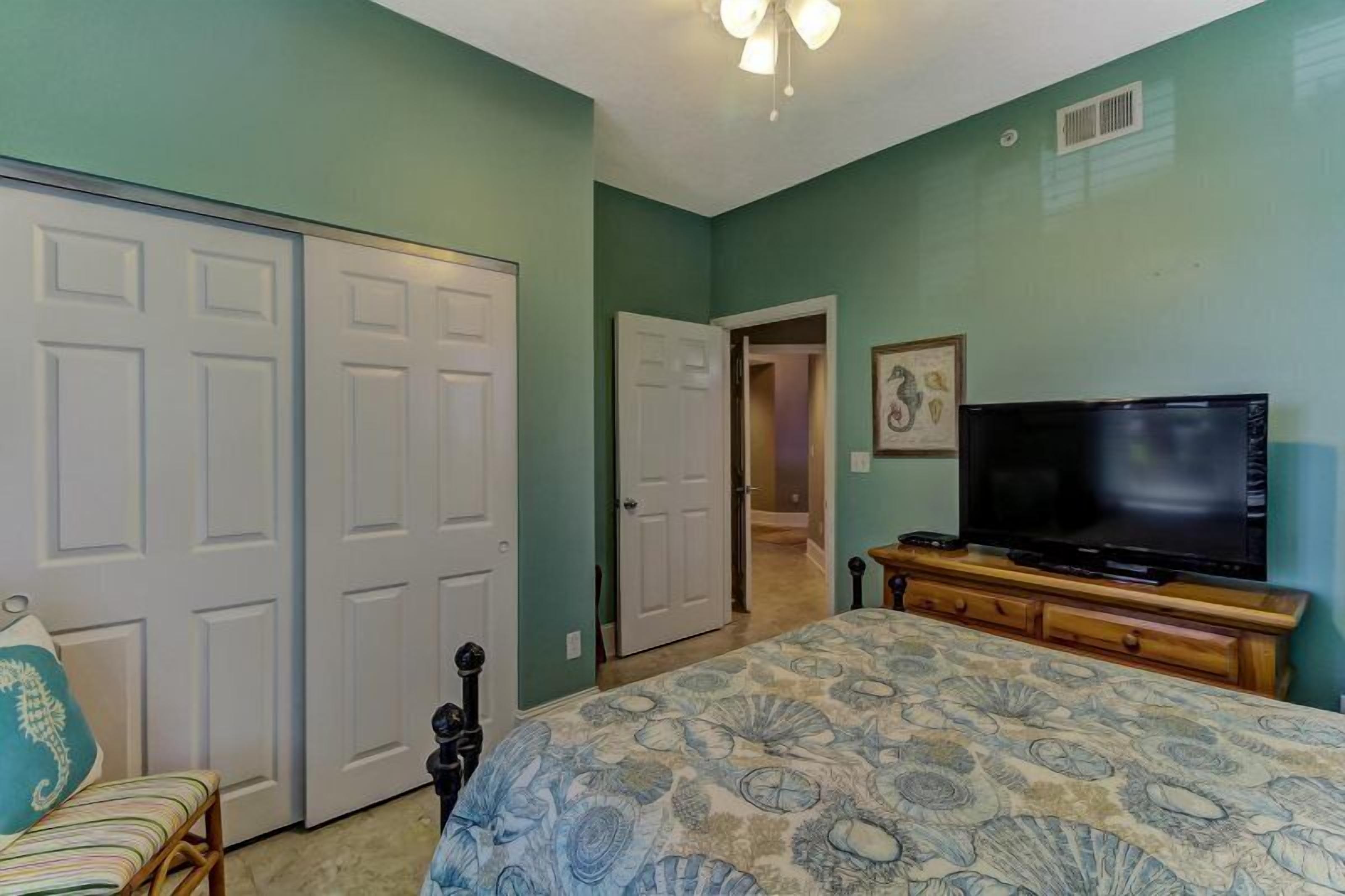 A bedroom with teal walls, a bed with a floral bedspread, a television on a wooden stand, and a door leading to another room.