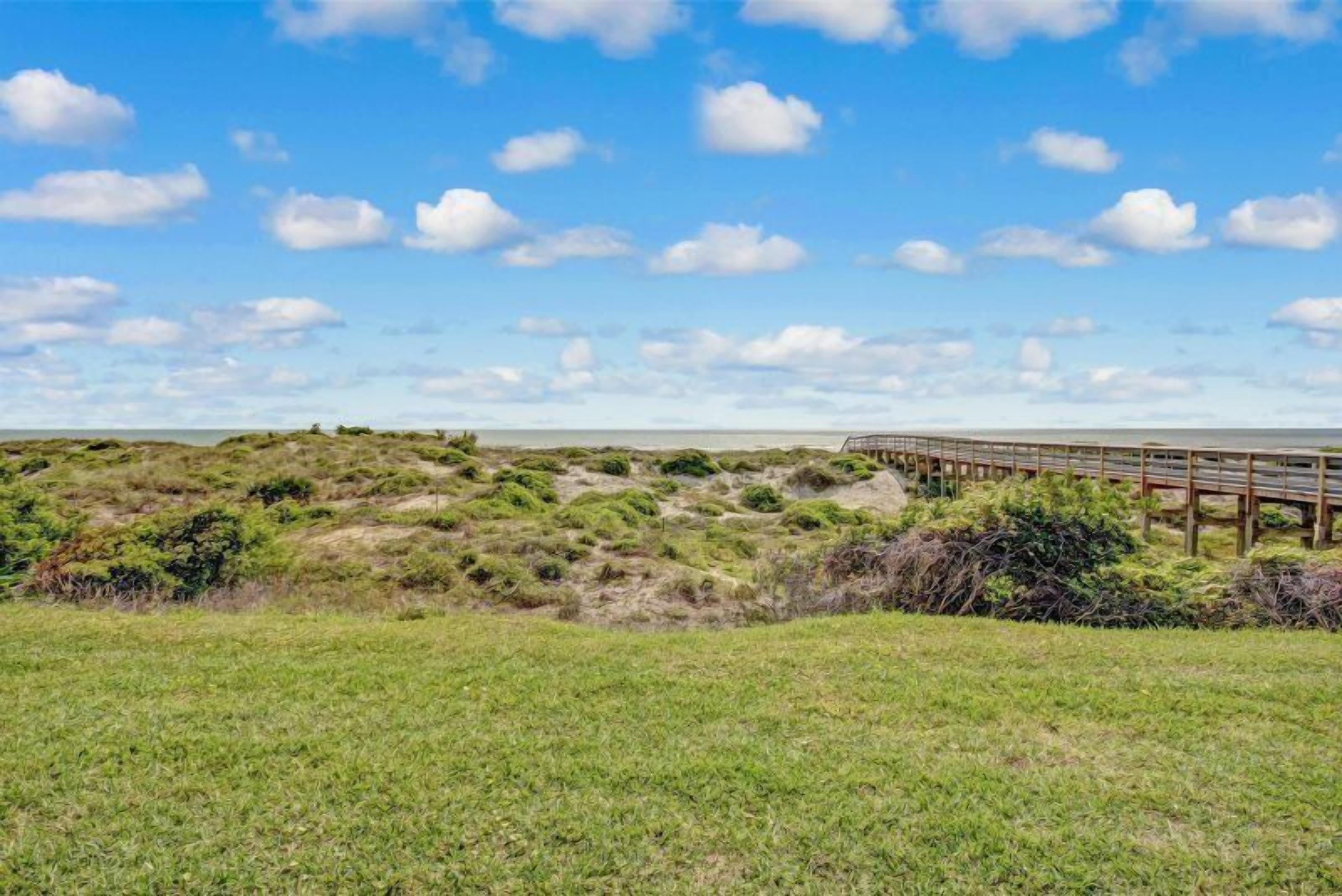 A coastal landscape with a wooden pier extending into the sea, surrounded by grassy dunes and sparse vegetation.