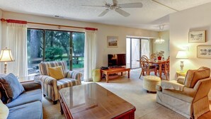A spacious living room with a large TV, comfortable seating, a ceiling fan, and a view through the glass doors. - 2135-beachwood (Fernandina Beach)