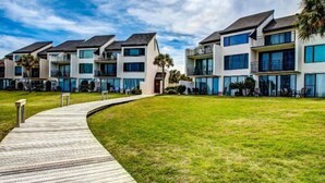 A modern apartment complex with a wooden boardwalk leading to the entrance, surrounded by well-maintained grass and palm trees.