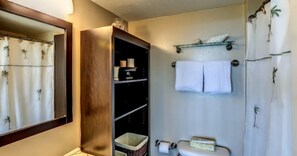 A bathroom with a wooden vanity, shelves with toiletries, a towel rack, and a shower curtain with palm tree patterns. - 1140-beach-walker (Fernandina Beach)