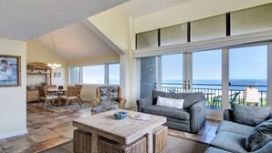 A spacious living area with a large window overlooking the ocean, a dining area with a table and chairs, and a cozy seating area with a sofa and a coffee table.