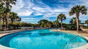 A swimming pool with a curved edge, surrounded by palm trees and lounge chairs, with a building in the background.