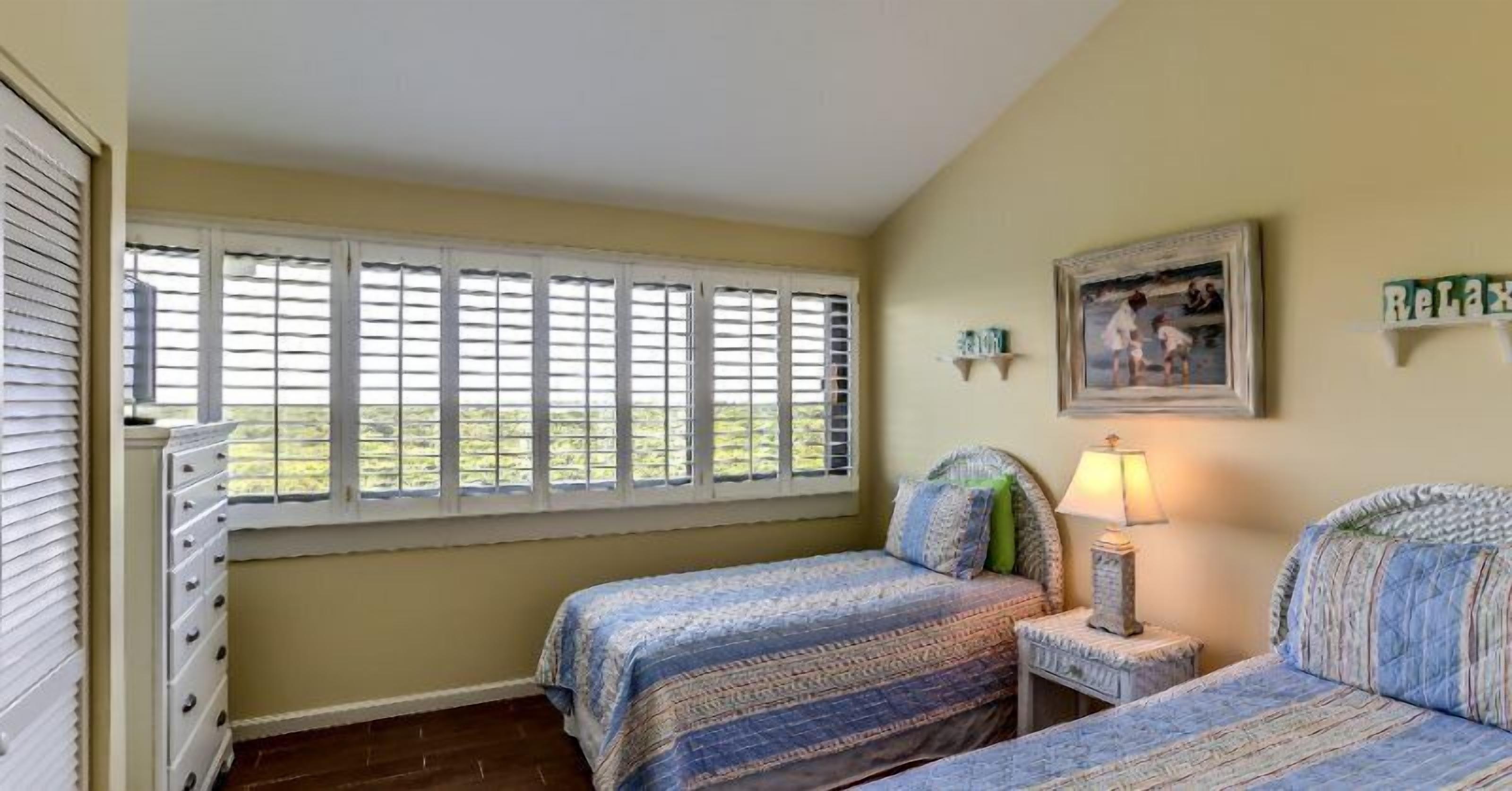 A bedroom with two beds, a dresser, a window with blinds, a framed picture on the wall, and a bedside lamp.