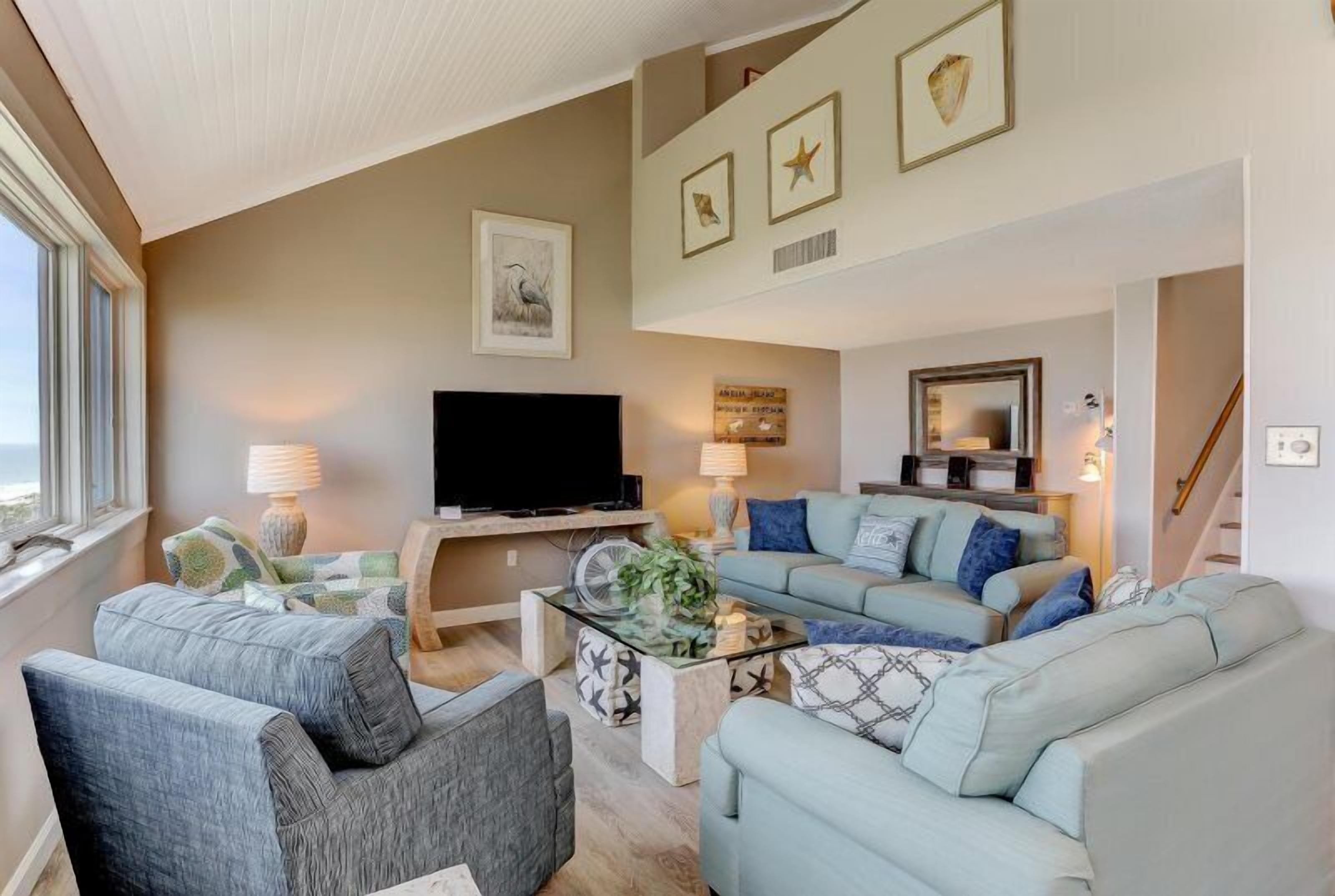 A living room with a sofa, armchairs, a coffee table, and a TV. There are framed pictures on the wall and a window with a view.
