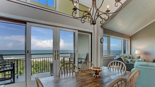 A dining area with a wooden table and chairs, a chandelier, and a view of the ocean through large sliding glass doors. - 1114-beach-walker (Fernandina Beach)