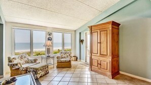 Habitación iluminada por el sol con un ventanal que da a una playa, un armario de madera y un sofá floral.
