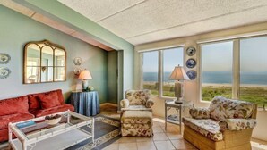 Una sala de estar con un sofá rojo, sillones con estampado floral, una mesa de centro y un amplio ventanal con vistas a la playa.