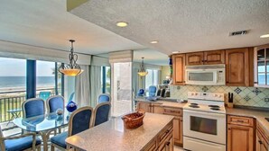 A kitchen with a large island, wooden cabinets, a microwave, and an oven. There is a dining area with chairs and a table, and large windows with a view. - 1182-beach-walker (Fernandina Beach)