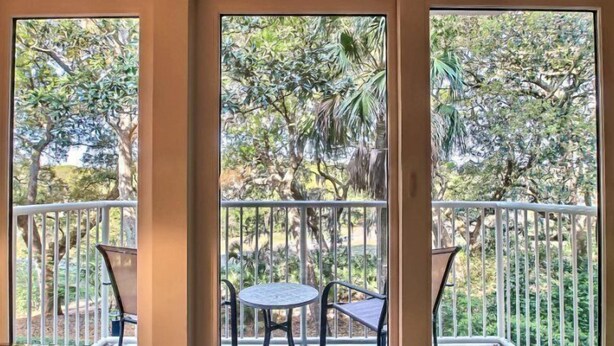 Un balcó amb taula i cadires, envoltat d'una barana metàl·lica, amb vista a un jardí exuberant ple d'arbres i plantes diverses.