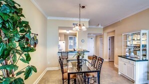 A dining area with a glass table and chairs, a kitchen with white cabinets, and a large green plant on the left. - 59-ocean-place (Fernandina Beach)