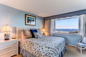 A bedroom with a bed, two bedside tables with lamps, a chair, a window with a view of the sea, and a framed picture on the wall.