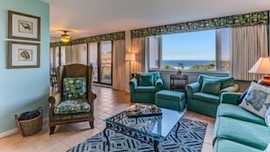 A living room with a view of the ocean, green sofas, a patterned coffee table, and a wicker basket.